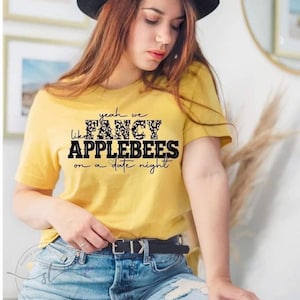 May include: A yellow t-shirt with black text that reads "yeah we like FANCY APPLEBEES on a date night". The text is in a leopard print font. The shirt is being worn by a person with brown hair and a black hat.