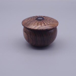 May include: A small, round, dark brown wooden lidded container with a visible wood grain pattern. The lid features a carved sunburst design radiating from a smooth, circular black centre inlay, creating a textured and detailed appearance.