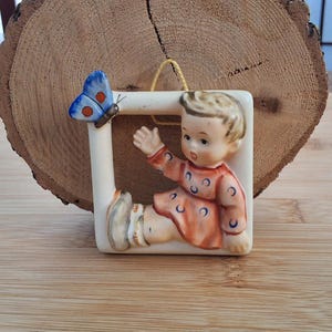Peut inclure: Une figurine en porcelaine d'un enfant en robe rouge à pois bleus tendant la main vers un papillon bleu. La figurine est encadrée dans un carré blanc.