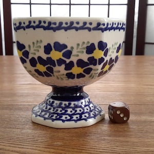 Peut inclure: Un bol en céramique blanche avec un motif floral bleu et jaune. Le bol est sur un piédestal et a une forme hexagonale. Un dé brun est à côté du bol.