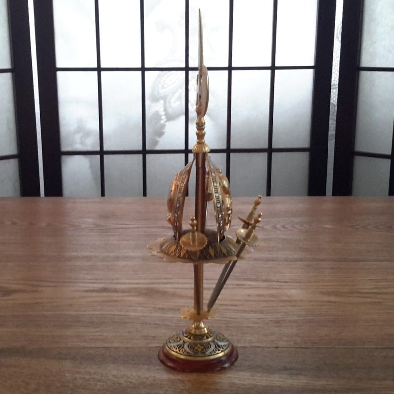 May include: A gold-colored metal decorative stand with a round top and three legs. The stand features a crown-like design with a crescent moon on top. There are two small swords in the stand.