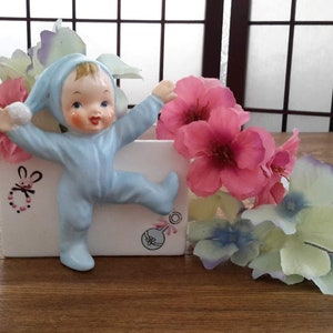 Peut inclure: Un pot en céramique blanc avec une figurine de bébé bleu grimpant sur le côté. Le pot est décoré d'un lapin rose et d'un hochet. Le bébé porte une gigoteuse bleue et un bonnet bleu. Le pot est entouré de fleurs roses et blanches.