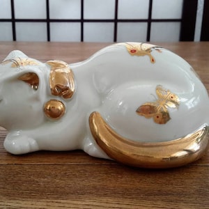 Peut inclure: Une figurine de chat endormi en porcelaine blanche avec des accents dorés. Le chat a un papillon doré sur le dos et une clochette dorée autour du cou.