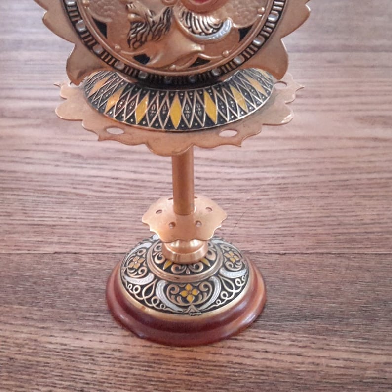 May include: A gold metal stand with a round top and three legs. The top features a decorative design with a central image of a winged figure with a halo. The stand has a brown wooden base.