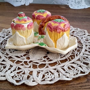 Peut inclure: Un ensemble de trois contenants à condiments en céramique en forme de fleurs stylisées avec des accents jaunes, verts et roses. Les contenants sont sur une dentelle blanche.