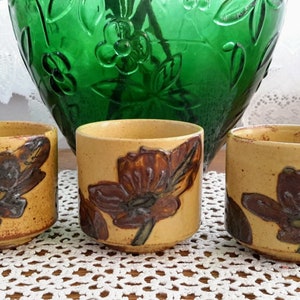 Peut inclure: Trois petites tasses en céramique avec des motifs floraux marrons sur fond jaune. Les tasses sont posées sur une dentelle blanche crochetée.