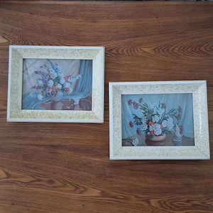 Peut inclure: Deux peintures de natures mortes florales encadrées avec des cadres blancs et un fond en bois clair. Les peintures représentent des fleurs blanches et roses dans des vases.