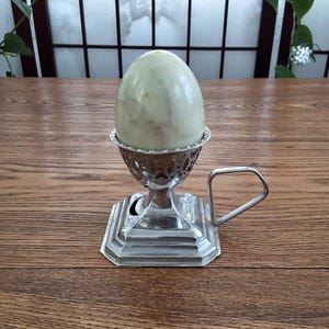 May include: A pale green, marble-effect egg sits in a silver egg cup with a handle. The egg cup has a decorative, openwork design and sits on a square, octagonal base. The egg is smooth and oval-shaped.