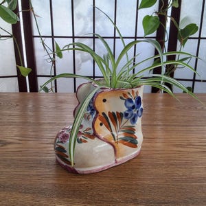 Peut inclure: Un pot de fleurs en céramique en forme de botte, de couleur crème, orné de motifs floraux et d'oiseaux bleus, oranges et verts. Il contient une plante araignée verte et blanche. La botte a une bordure rouge et repose sur une surface en bois.