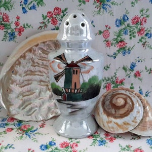 Peut inclure: Un saupoudreur en céramique blanche avec un moulin à vent peint dessus. Le saupoudreur est posé sur une nappe à fleurs avec deux coquillages.