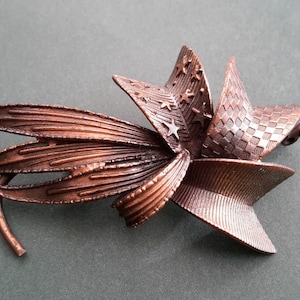 May include: A copper-toned metal brooch in the shape of a stylised flower with intricate details. The flower has multiple petals with a textured surface and a star-shaped centre.