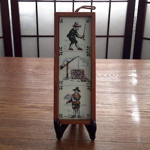 May include: A wooden framed tile picture with three scenes of people in traditional clothing. The top scene shows a person swinging a stick, the middle scene shows a person drawing water from a well, and the bottom scene shows a person holding a piece of paper.
