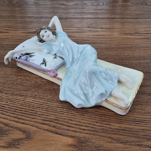 May include: A porcelain figurine of a woman reclining on a bed-shaped base. The woman wears a light blue gown and rests on a pillow with floral designs. The base is cream-coloured with a gold trim.