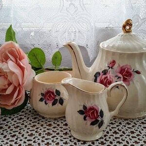Peut inclure: Un service à thé vintage comprenant une théière, un pot à crème et un sucrier, tous ornés de motifs de roses roses. L'ensemble est de couleur crème avec des bordures dorées. Des roses roses fraîches et un rideau en dentelle complètent la scène.