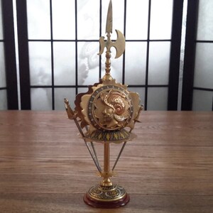 May include: A gold-toned metal decorative stand with a round top and three legs. The stand features a shield with a knight's head and a spear with an axe on top.