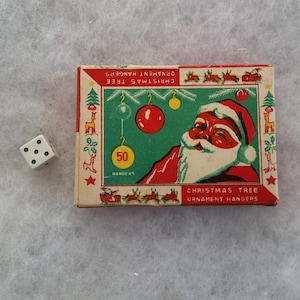 May include: A vintage box of Christmas tree ornament hangers with a Santa Claus illustration. The box is red, green, and white, with the text "CHRISTMAS TREE ORNAMENT HANGERS". A white die with black dots is next to the box.