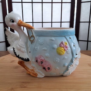 Peut inclure: Un pot de fleurs en céramique blanche en forme de cigogne avec un motif de couche bleue. La couche a un hochet jaune, un nœud rose et des blocs qui disent "A, P, 1, B".