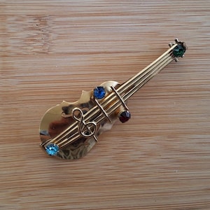 Peut inclure: Une broche en forme de violon dorée avec des strass multicolores. La broche représente une clé de sol et des cordes.