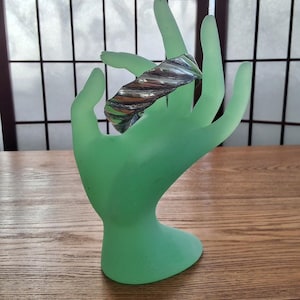 May include: A silver and copper bracelet with a textured, wavy design. The bracelet is displayed on a green hand-shaped stand.