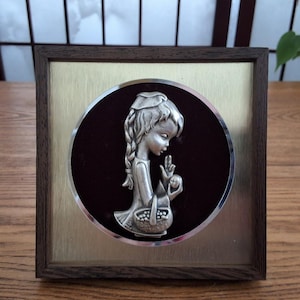May include: A framed silver relief sculpture of a young girl holding a basket of fruit. The sculpture is set against a dark red circular background, framed by a gold-colored metal and a dark brown wooden frame. The girl is wearing a hat and has long braids.