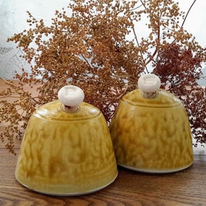 May include: Two ceramic bell-shaped containers with white lids. The containers are a yellow-brown color with a mottled glaze. They are sitting on a wooden surface.