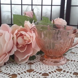 Peut inclure: Un pot à crème en verre rose avec un motif floral posé sur un napperon blanc en crochet. Le pot à crème a une anse et un pied. Le pot à crème est entouré de roses roses.