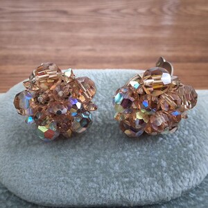 Peut inclure: Une paire de boucles d'oreilles vintage à clip. Chaque boucle d'oreille présente un groupe de perles facettées et irisées dans des tons ambre et marron. Les perles sont disposées en forme de fleur, avec un clip argenté au dos.