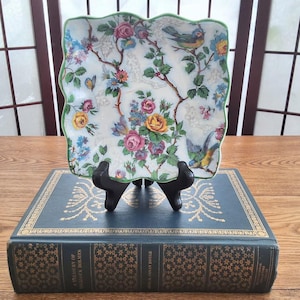 May include: A square decorative plate with a floral and bird design, featuring pink, yellow, and blue flowers, and perched birds on branches. The plate has a scalloped green edge and is displayed on a black stand.