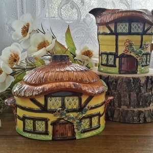Peut inclure: Un ensemble de deux pièces de cuisine en céramique en forme de cottage. La plus grande pièce est un pichet avec un toit de chaume brun et une façade jaune. La plus petite pièce est un sucrier avec un toit de chaume brun et une façade jaune. Les deux pièces ont des bordures brunes et des fenêtres.