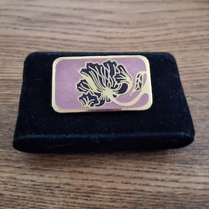 Peut inclure: Une broche rectangulaire avec une bordure dorée et un fond mauve. La broche présente un motif floral noir. La broche est présentée sur une surface de velours noir.