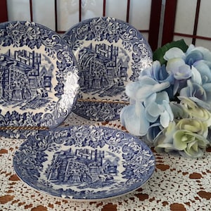 Peut inclure: Trois assiettes en céramique bleu et blanc avec un motif floral et une scène d'un bâtiment avec un moulin à vent. Les assiettes sont posées sur une nappe en dentelle blanche.