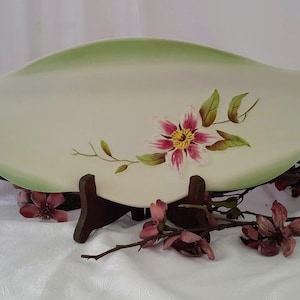 May include: A white oval serving dish with a green rim and a hand-painted pink flower with yellow center. The dish is on a wooden stand.