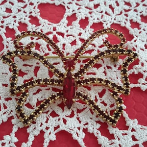 Peut inclure: Une broche en forme de papillon en or avec des accents de strass rouges. Le papillon a un grand strass rouge au centre.
