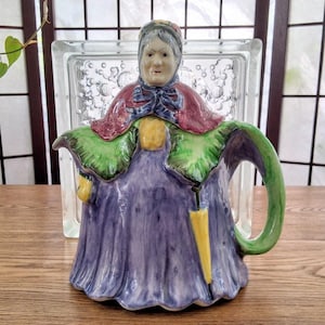 May include: A ceramic teapot shaped like an elderly person wearing a purple dress, a maroon cape, and a green shawl. The figure holds a yellow umbrella, and the teapot features a green handle and a spout integrated into the purple dress design.