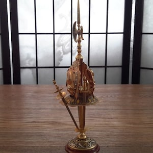 May include: A gold-colored decorative stand with a round top and three legs. The stand features a sword, a spear, and a shield.
