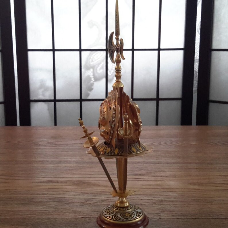 May include: A gold-colored decorative stand with a round top and three legs. The stand features a sword, a spear, and a shield.