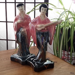 May include: Two ceramic figurines of women in traditional Japanese clothing. The woman on the left is holding a fan, and the woman on the right is playing a stringed instrument. Both figurines are painted in shades of pink, black, and white.
