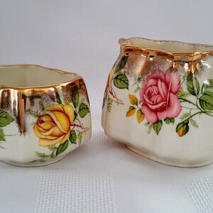 Peut inclure: Un ensemble sucrier et crémier vintage avec un motif floral. L'ensemble est blanc avec des accents dorés et présente des roses roses et jaunes.