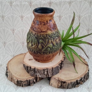 Peut inclure: Un vase en céramique brun et vert avec un motif en relief complexe posé sur trois tranches de bois. Le vase a un corps arrondi et une ouverture étroite.