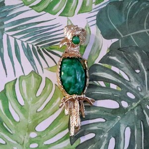 Peut inclure: Broche perroquet dorée avec un grand corps en pierre verte ovale. L'oiseau a une crête, un bec et des plumes de queue détaillés. Une petite pierre verte orne la tête. La broche est posée sur un fond de feuilles tropicales.