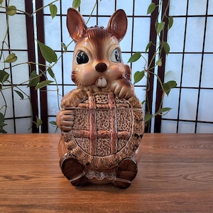May include: A ceramic squirrel cookie jar with a basket design. The squirrel is brown and tan with large eyes and a cute expression. The basket is a lighter brown with a woven pattern. The jar is sitting on a wooden surface.