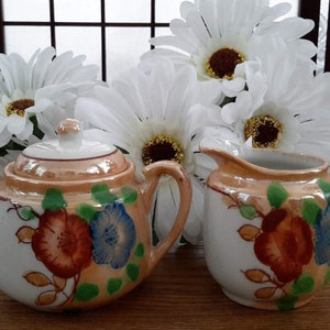 Peut inclure: Un ensemble sucrier et crémier en porcelaine vintage avec des motifs floraux dans des tons d'orange, bleu et vert. L'ensemble est orné d'une bordure dorée et d'un motif floral.