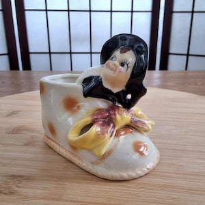 May include: A white ceramic planter shaped like a shoe with brown polka dots and a yellow bow. A small figurine of a woman wearing a black hat and dress is sitting on top of the shoe.