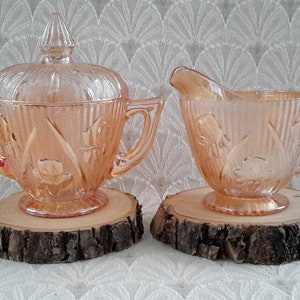 Peut inclure: Un ensemble vintage de pot à crème et sucrier en verre rose avec un motif floral. L'ensemble est posé sur deux sous-verres en bois.