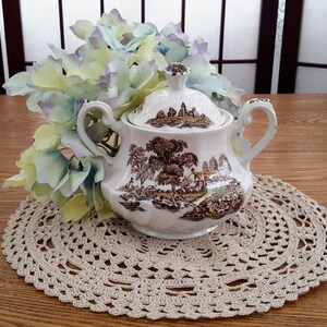 Peut inclure: Sucrier en porcelaine blanche avec un couvercle, représentant une scène de campagne brune et verte avec une maison et des arbres. Le bol a deux anses et est posé sur un napperon en crochet.