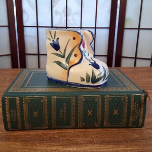 May include: A ceramic boot-shaped vase with a cream base, decorated with blue and red floral designs and green leaves. The vase rests on a vintage green book with gold lettering.
