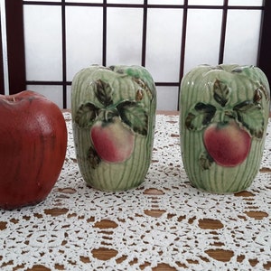 Peut inclure: Deux salières et poivrières en céramique verte avec des motifs de pommes en relief. Les salières sont sur une nappe en dentelle blanche.