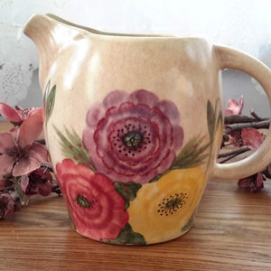 May include: A cream-coloured ceramic pitcher with a floral design. The pitcher features a yellow, red, and purple flower design with green leaves.