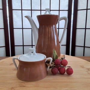 Peut inclure: Un ensemble de cafetière et de sucrier en céramique marron et blanc. La cafetière a un couvercle blanc et un corps marron avec un bec et une poignée blancs. Le sucrier a un couvercle blanc et un corps marron avec deux poignées blanches.