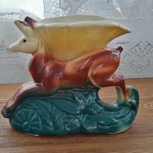 May include: A ceramic vase shaped like a deer with a yellow and white leaf on its back. The deer is standing on a green base with floral details.